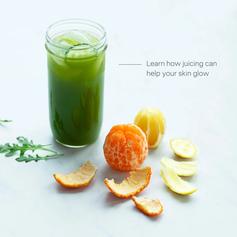 A glass of Skin Health green juice with cucumber slices, surrounded by peeled citrus fruit, citrus peels, and arugula, sits on a light surface alongside the text: Discover how Skin Health juicing can help your skin glow.