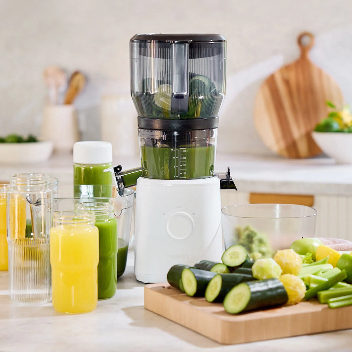 A juicer with green juice inside sits on a kitchen counter surrounded by glasses of yellow and green juice, sliced cucumbers, lemons, celery, and a cutting board in the background—a perfect setup for batch juicing with the Large Hopper.