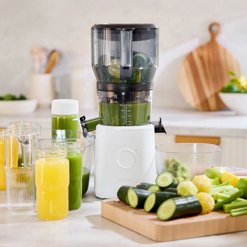 A juicer with green juice inside sits on a kitchen counter surrounded by glasses of yellow and green juice, sliced cucumbers, lemons, celery, and a cutting board in the background—a perfect setup for batch juicing with the Large Hopper.