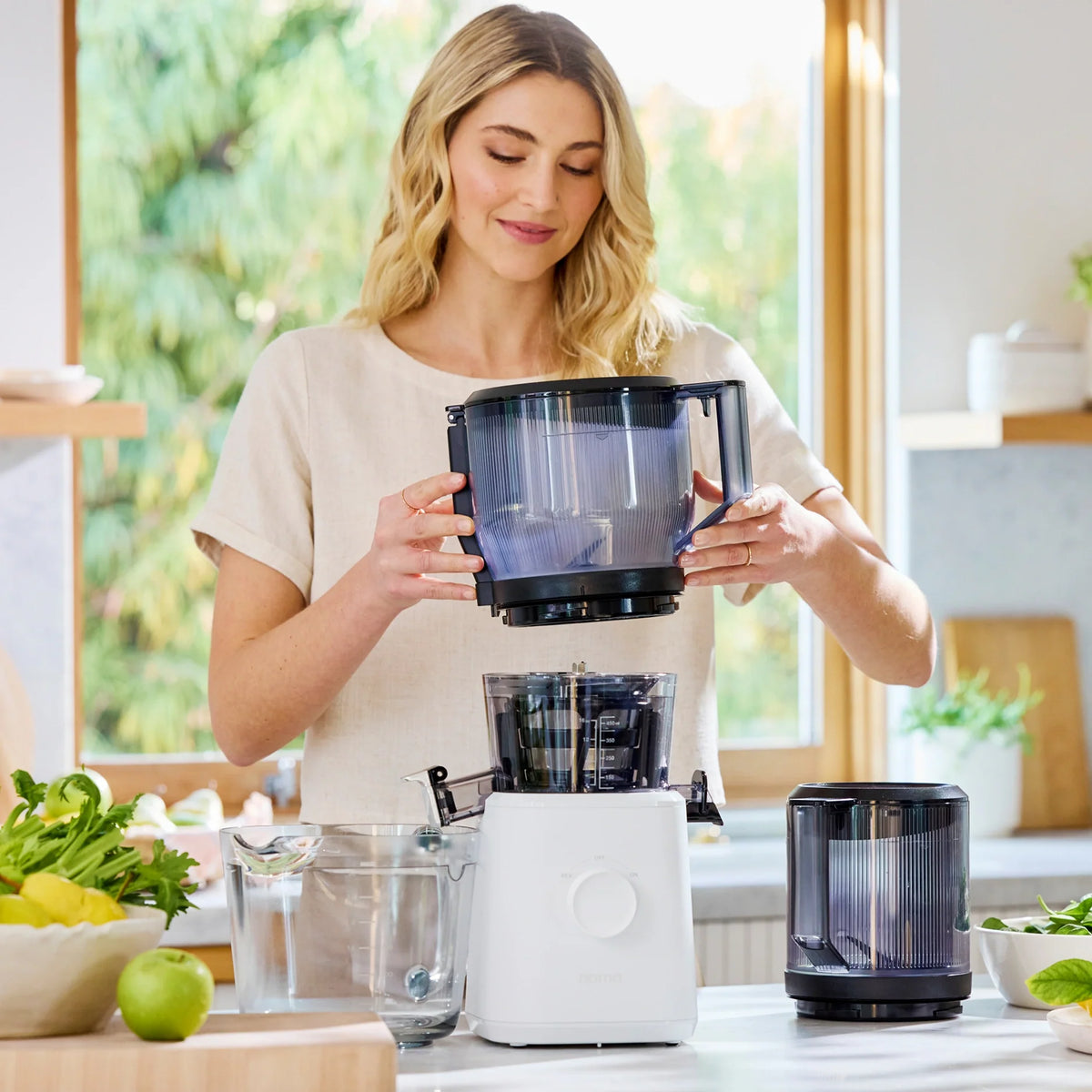 In a bright kitchen filled with fresh vegetables and lemons, a woman assembles the Large Hopper food processor with its juicer attachment, while sunlight streams through a window overlooking lush greenery—ideal for convenient batch juicing.