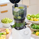 A juicer filled with green apples and celery sits on a kitchen counter surrounded by fresh fruits and vegetables—including grapes, pears, and spinach—making it perfect for batch juicing with the Large Hopper or your favorite juicer attachment.
