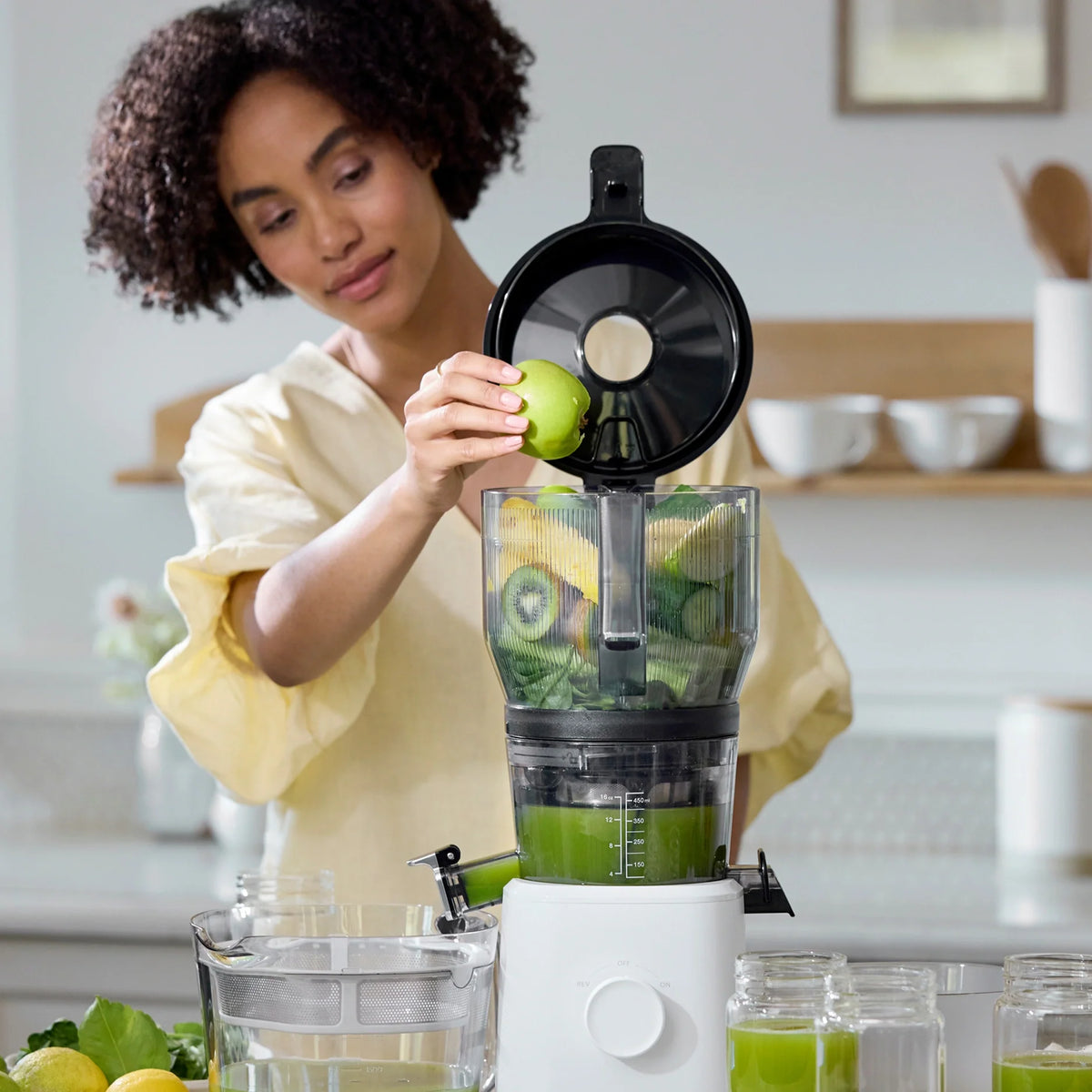 A woman places a green apple into the Large Hopper, using its innovative juicer attachment to simplify batch juicing in a bright kitchen. Glasses of green juice and fresh produce sit on the counter nearby.