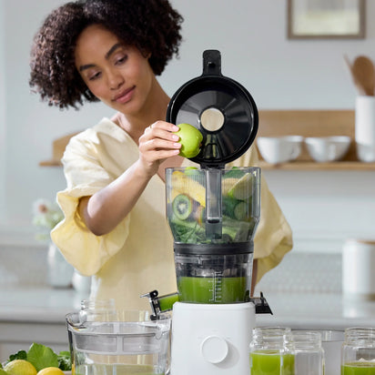 A woman places a green apple into the Large Hopper, using its innovative juicer attachment to simplify batch juicing in a bright kitchen. Glasses of green juice and fresh produce sit on the counter nearby.