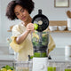 A woman places a green apple into the Large Hopper, using its innovative juicer attachment to simplify batch juicing in a bright kitchen. Glasses of green juice and fresh produce sit on the counter nearby.