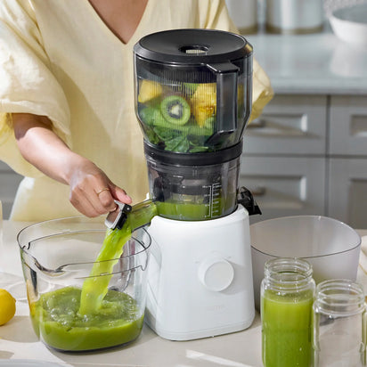 Wearing a light yellow shirt, a person uses the Large Hopper with its juicer attachment to extract green juice from leafy greens, kiwi, and lemon. Juice flows into a pitcher as part of their batch juicing process, with empty glass jars arranged on the kitchen counter.