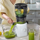 Wearing a light yellow shirt, a person uses the Large Hopper with its juicer attachment to extract green juice from leafy greens, kiwi, and lemon. Juice flows into a pitcher as part of their batch juicing process, with empty glass jars arranged on the kitchen counter.