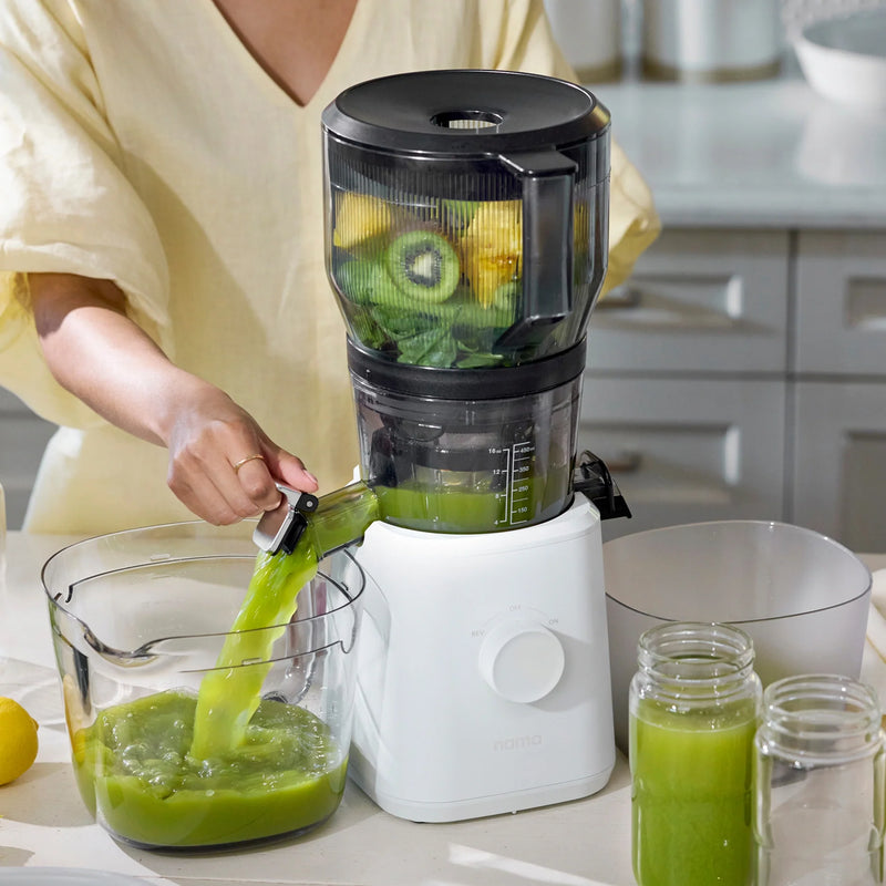 Wearing a light yellow shirt, a person uses the Large Hopper with its juicer attachment to extract green juice from leafy greens, kiwi, and lemon. Juice flows into a pitcher as part of their batch juicing process, with empty glass jars arranged on the kitchen counter.