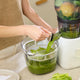 A person operates a juicer, pouring fresh green juice into a Large Pitcher Set on a kitchen counter. Nearby, various accessories like the included strainer and pitcher from the Large Pitcher Set are arranged for convenient juice storage. Celery and leafy greens are visible in the juicer.