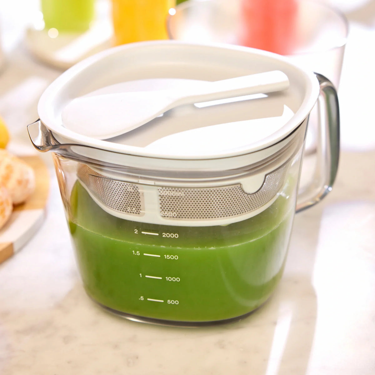 A Large Pitcher Set filled with green liquid, complete with a white strainer and spatula, sits on a kitchen counter—an ideal setup for those seeking convenient juice storage or using juicer accessories.