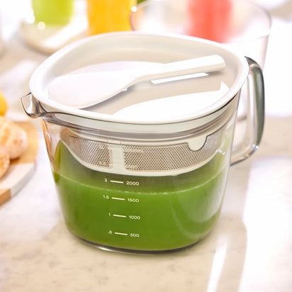 A Large Pitcher Set filled with green liquid, complete with a white strainer and spatula, sits on a kitchen counter—an ideal setup for those seeking convenient juice storage or using juicer accessories.