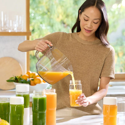 A woman pours orange juice from the Large Pitcher Set, which includes a strainer, into a glass in her kitchen, with several bottles of green and orange juice—perfect for convenient juice storage—arranged neatly on the counter.