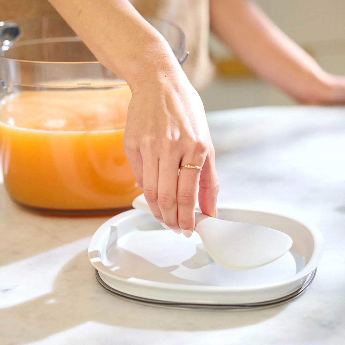 A hand places a white spoon on a white divided plate on a marble counter, with the Large Pitcher Set—containing orange liquid and juicer accessories like a pitcher and strainer—visible in the background.