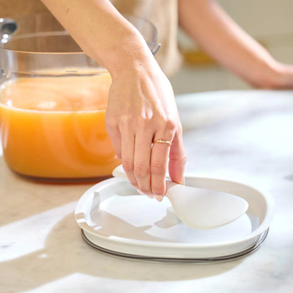 A hand places a white spoon on a white divided plate on a marble counter, with the Large Pitcher Set—containing orange liquid and juicer accessories like a pitcher and strainer—visible in the background.