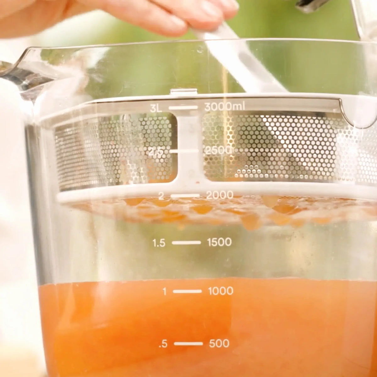 Close-up of the Large Pitcher Set filled with orange liquid up to approximately 1.5 liters, showcasing built-in pitcher and strainer juicer accessories, with a hand holding a spoon, possibly stirring or mixing the contents.