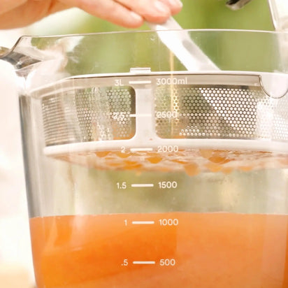 Close-up of the Large Pitcher Set filled with orange liquid up to approximately 1.5 liters, showcasing built-in pitcher and strainer juicer accessories, with a hand holding a spoon, possibly stirring or mixing the contents.