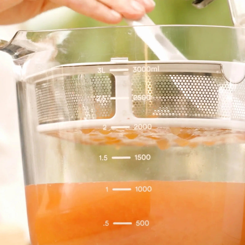 Close-up of the Large Pitcher Set filled with orange liquid up to approximately 1.5 liters, showcasing built-in pitcher and strainer juicer accessories, with a hand holding a spoon, possibly stirring or mixing the contents.