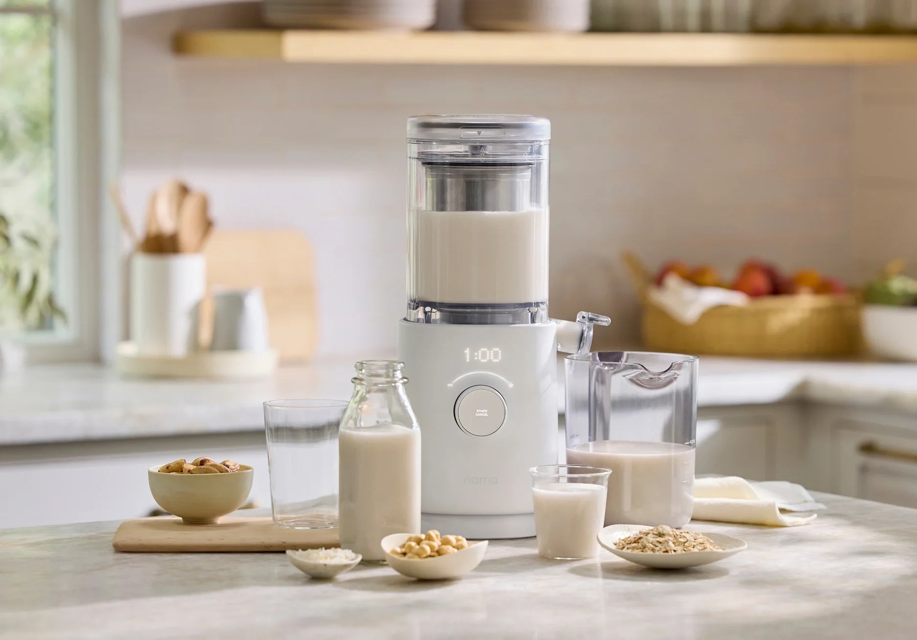 A nut milk maker on a kitchen counter surrounded by bowls of nuts, a glass, a jar, a pitcher, and a small bowl of oats. The machine displays 1:00 and is filled with white liquid, likely plant milk.
