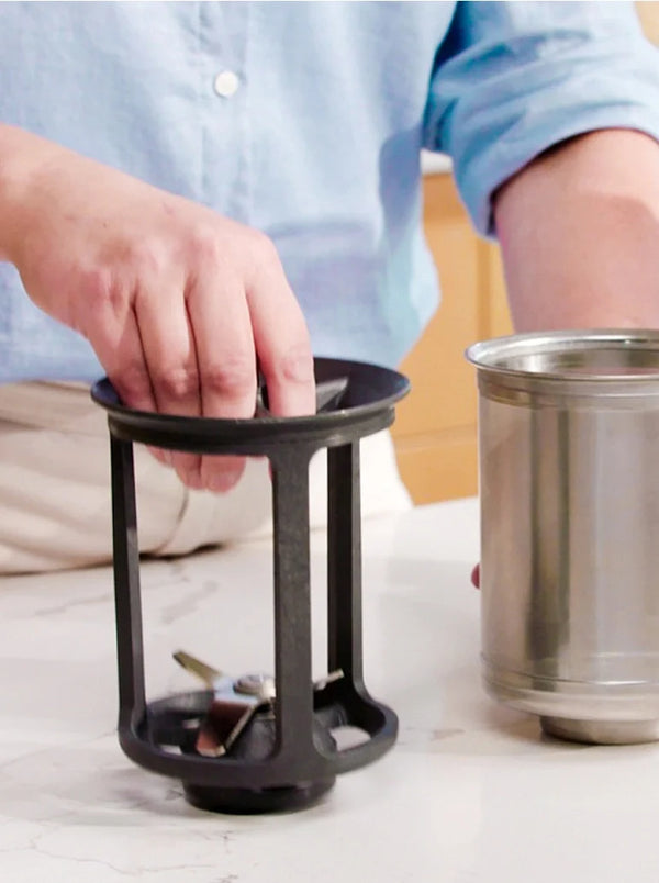 A person in a light blue shirt holds a black metal blender blade assembly next to a stainless steel blender container on a white countertop.
