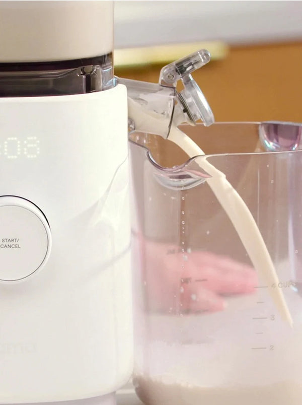 A white kitchen appliance dispenses a creamy liquid into a clear measuring cup. A hand is visible in the background, and the appliance has a visible Start/Cancel button.