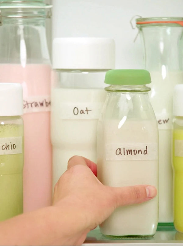 A hand reaching for a glass bottle labeled almond among other bottles of plant-based milk labeled oat, cashew, pistachio, and strawberry.