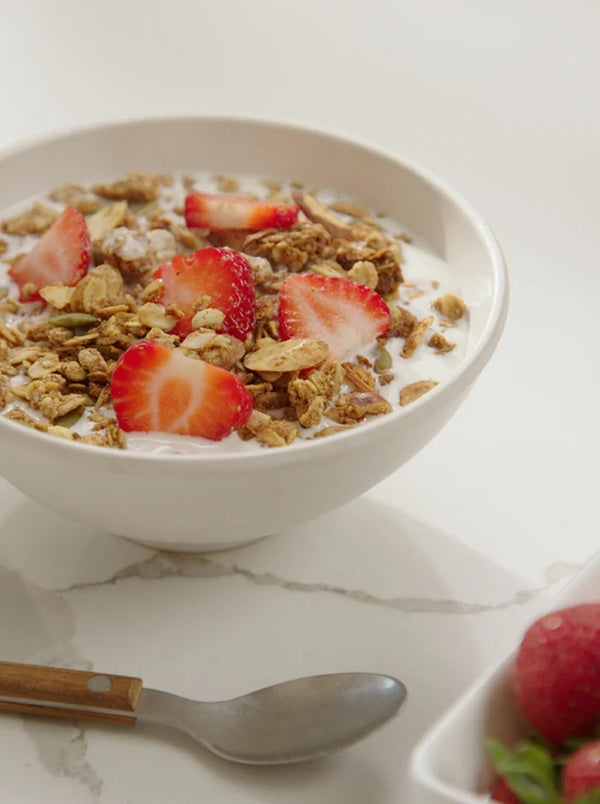 A white bowl filled with yogurt, granola, and sliced strawberries sits on a white surface next to a spoon and a partial view of another bowl of strawberries.