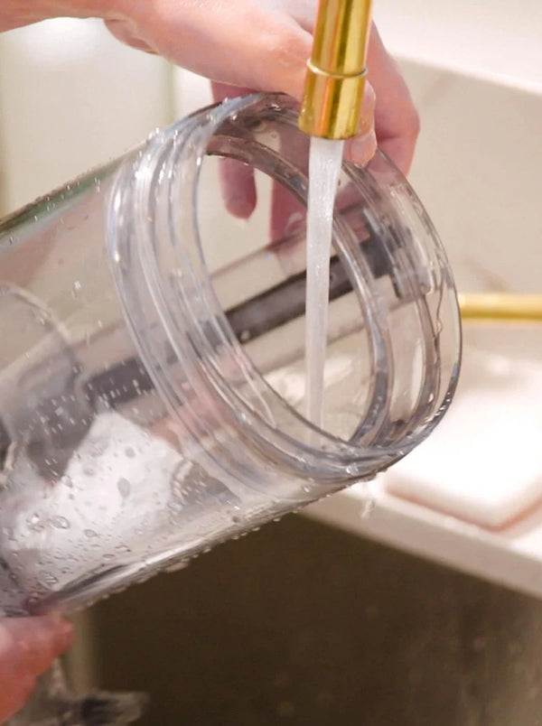 A person rinses a clear blender jar under running water from a gold faucet in a sink, with water and droplets visible inside and outside the jar.