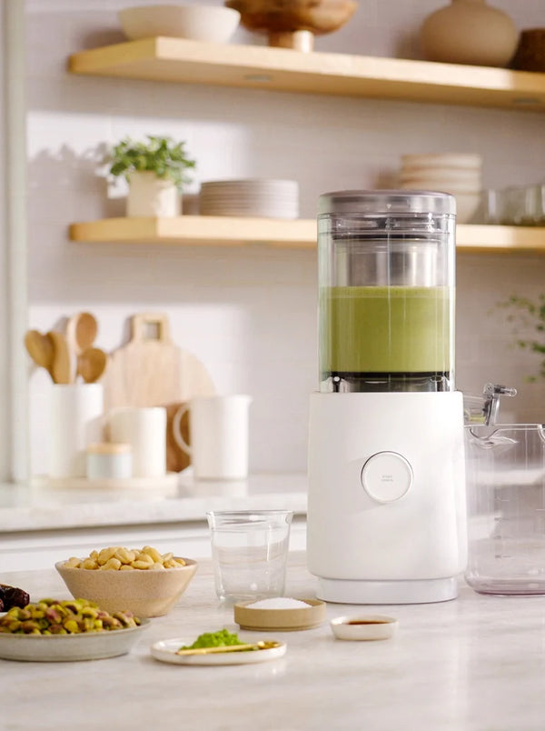 A white countertop juicer machine filled with green juice, shown in a bright, modern kitchen setting with bowls of ingredients and wooden shelves in the background.