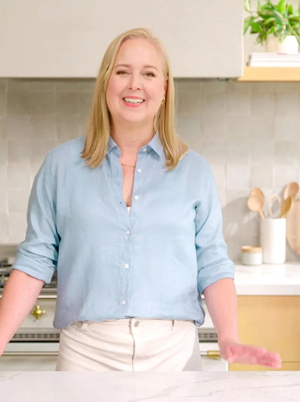 A smiling blonde woman wearing a light blue button-down shirt and cream pants, standing in a bright modern kitchen with white cabinets and a tiled backsplash.