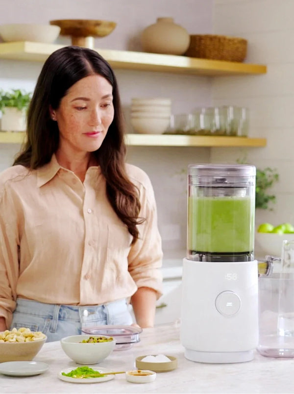 A woman in a beige blouse stands at a kitchen counter next to a white countertop blender filled with green juice, with bowls of ingredients and wooden shelves in the background.