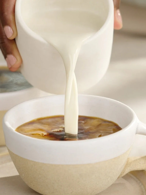 A hand pours creamy milk from a small white pitcher into a cup of coffee, creating swirling patterns on the coffees surface.