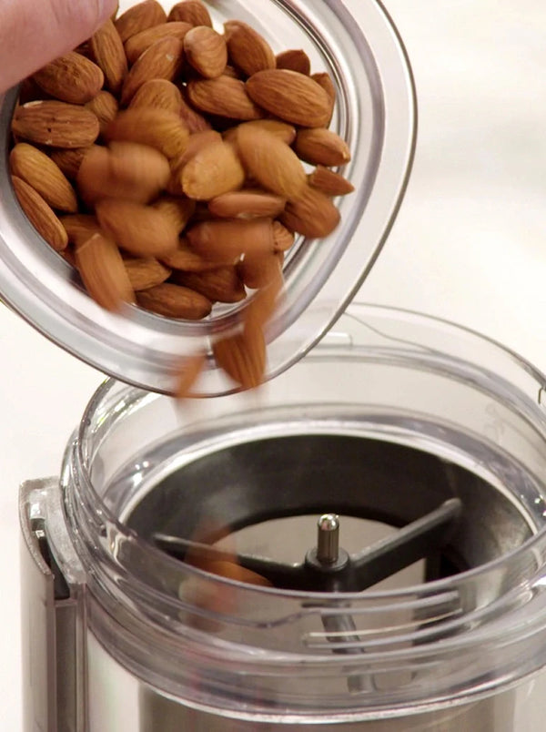 A person pours raw almonds from a glass bowl into an electric grinder or blender, preparing to blend the nuts.