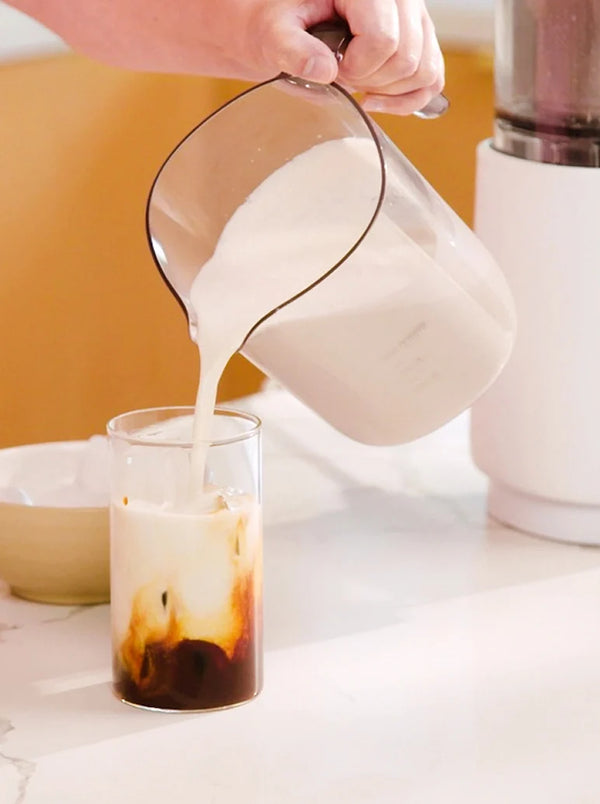 A hand pours creamy white liquid from a pitcher into a glass of iced coffee on a white countertop.