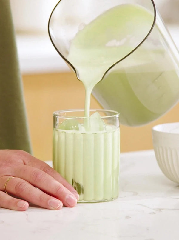 A hand holds a glass with ice as a pale green creamy drink is poured into it from a pitcher. The scene is set on a white countertop with a blurred background.