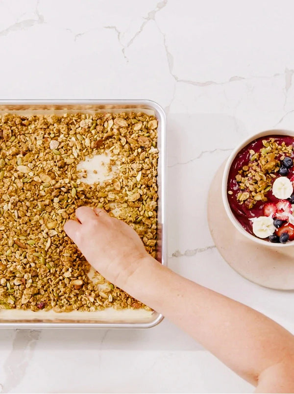 A hand sprinkles granola over a baking tray on a white counter. Next to it is a bowl of smoothie topped with bananas, blueberries, and granola.