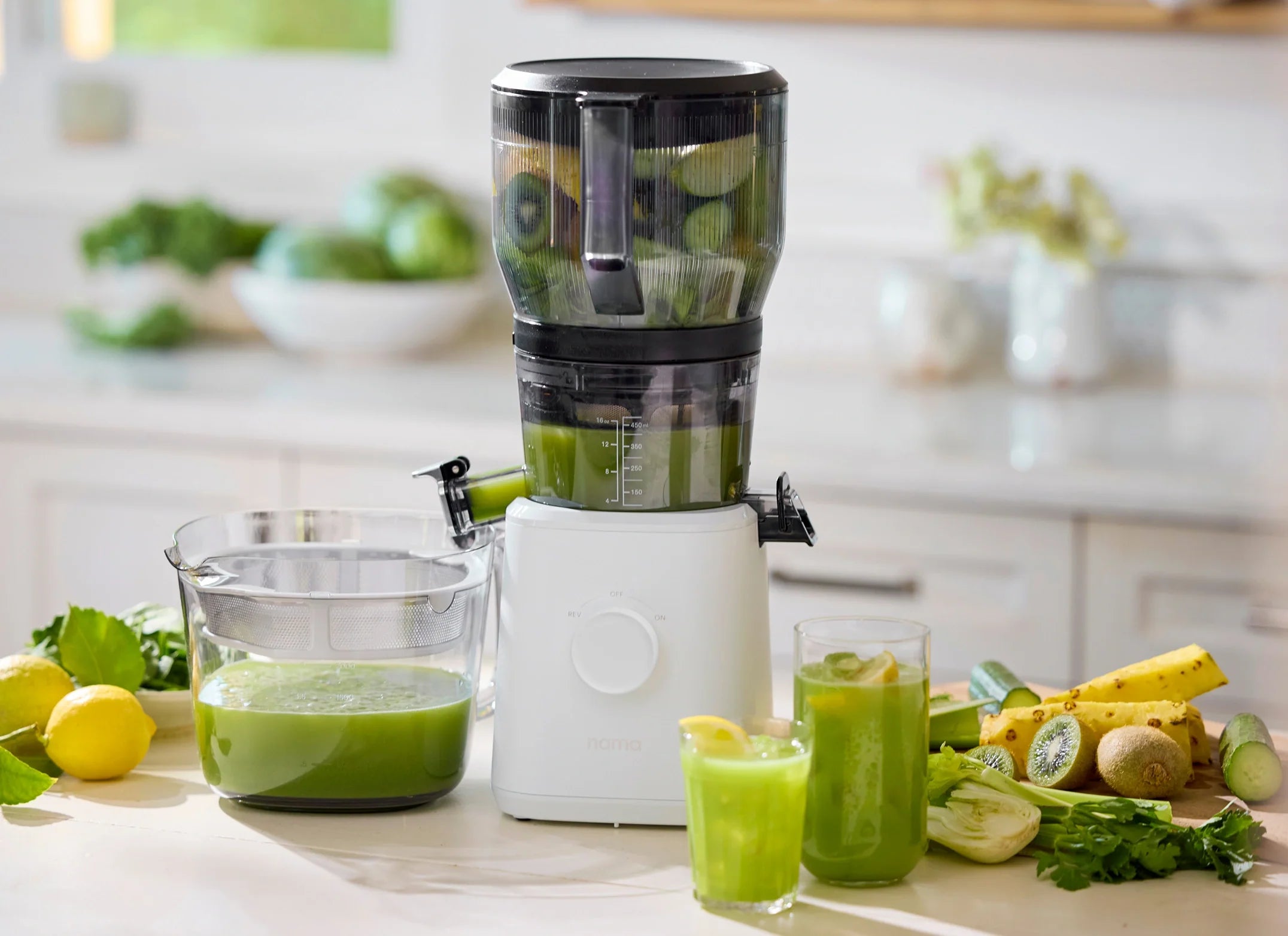 A juicer extracts green juice from fruits and vegetables on a kitchen counter, with a glass pitcher, two glasses of juice, and fresh produce like lemons, celery, and kiwi nearby. Bright, natural light fills the scene.