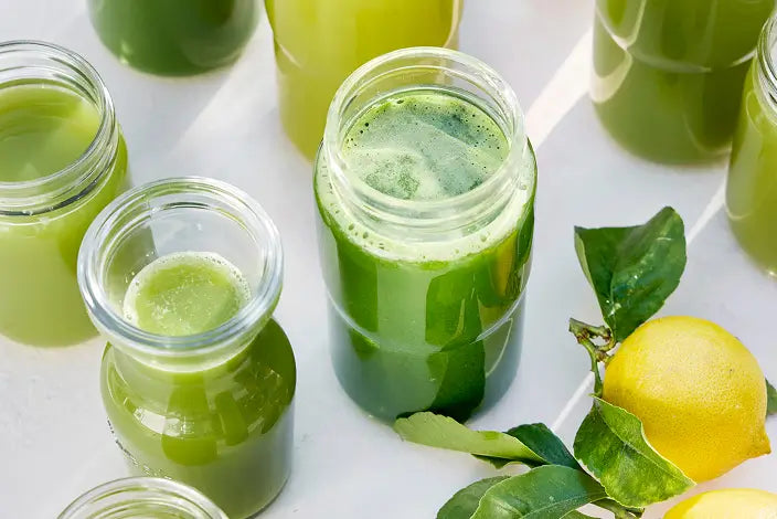 Several glass jars filled with green juice are arranged on a white surface, with fresh green leaves and a yellow lemon placed nearby.