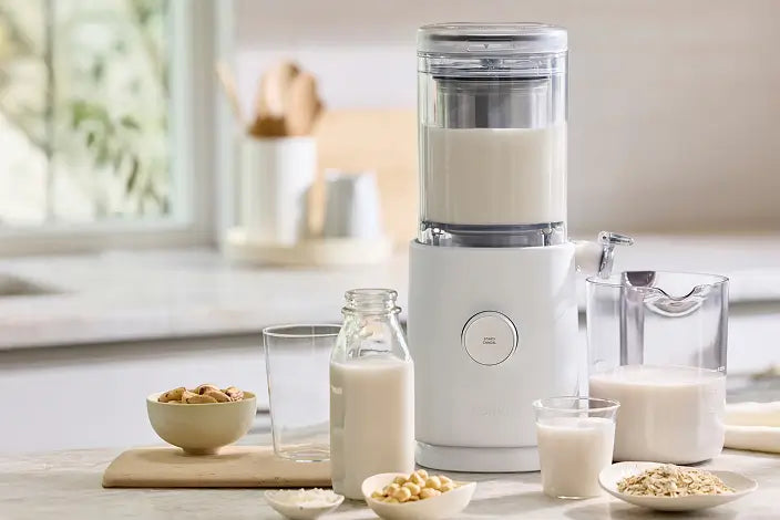 A nut milk maker sits on a kitchen counter, surrounded by a glass bottle, a glass, a pitcher of milk, and bowls of nuts and oats. The background is softly lit, with blurred kitchen utensils and a window.