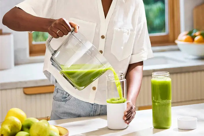 A person in a white shirt pours green juice from a pitcher into a glass on a kitchen counter. A tall jar of green juice and a bowl of apples are nearby.