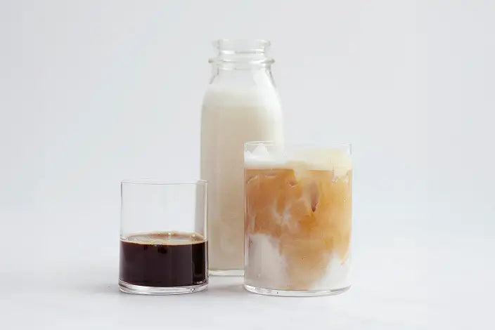 A bottle of milk, a glass of black coffee, and a glass of iced coffee with milk are arranged on a white surface with a plain background.
