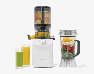 A white juicer and blender with fruits inside, next to two glasses of green and orange juice. The blender jar contains bananas, strawberries, spinach, and mango. The background is plain white.