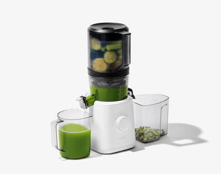 A juicer with green juice inside processes whole fruits and vegetables. A pitcher of green juice and a container of pulp are placed beside it, all on a white background.