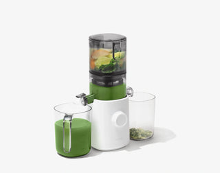 A white juicer with leafy greens and fruit inside, green juice pouring into a container on the left, and pulp collected in a container on the right, all set against a plain white background.