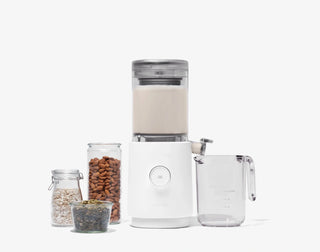 A white countertop appliance with a transparent top, surrounded by a measuring cup and jars filled with almonds, oats, and seeds, against a plain white background.