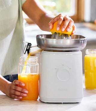 A person uses an electric citrus juicer to squeeze an orange, filling a glass with fresh orange juice. Another glass of juice sits nearby on the counter.