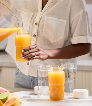 A person pours orange juice from a pitcher into a glass jar, with more empty jars and citrus fruits on a kitchen counter.