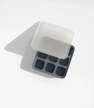 A black silicone ice cube tray with six square compartments, shown with a translucent plastic lid partially covering it, on a white background.