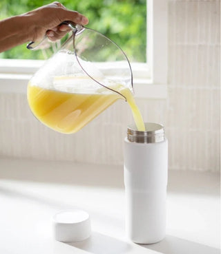 A hand pours yellow juice from a glass pitcher into a white insulated bottle on a white countertop, with a white lid nearby and greenery visible through a window in the background.