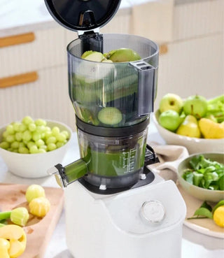 A juicer filled with green apples and grapes sits on a kitchen counter, surrounded by bowls of fresh green fruits.