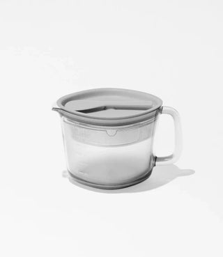 A transparent plastic measuring cup with a lid, containing white powder, sits on a white surface with a soft shadow cast to the right.