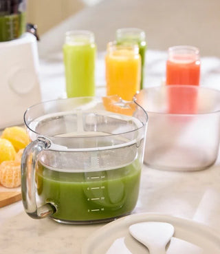 A glass measuring cup filled with green juice sits on a counter; behind it are jars of colorful juice—green, yellow, orange, and pink. Sliced citrus fruits and kitchen utensils are nearby.